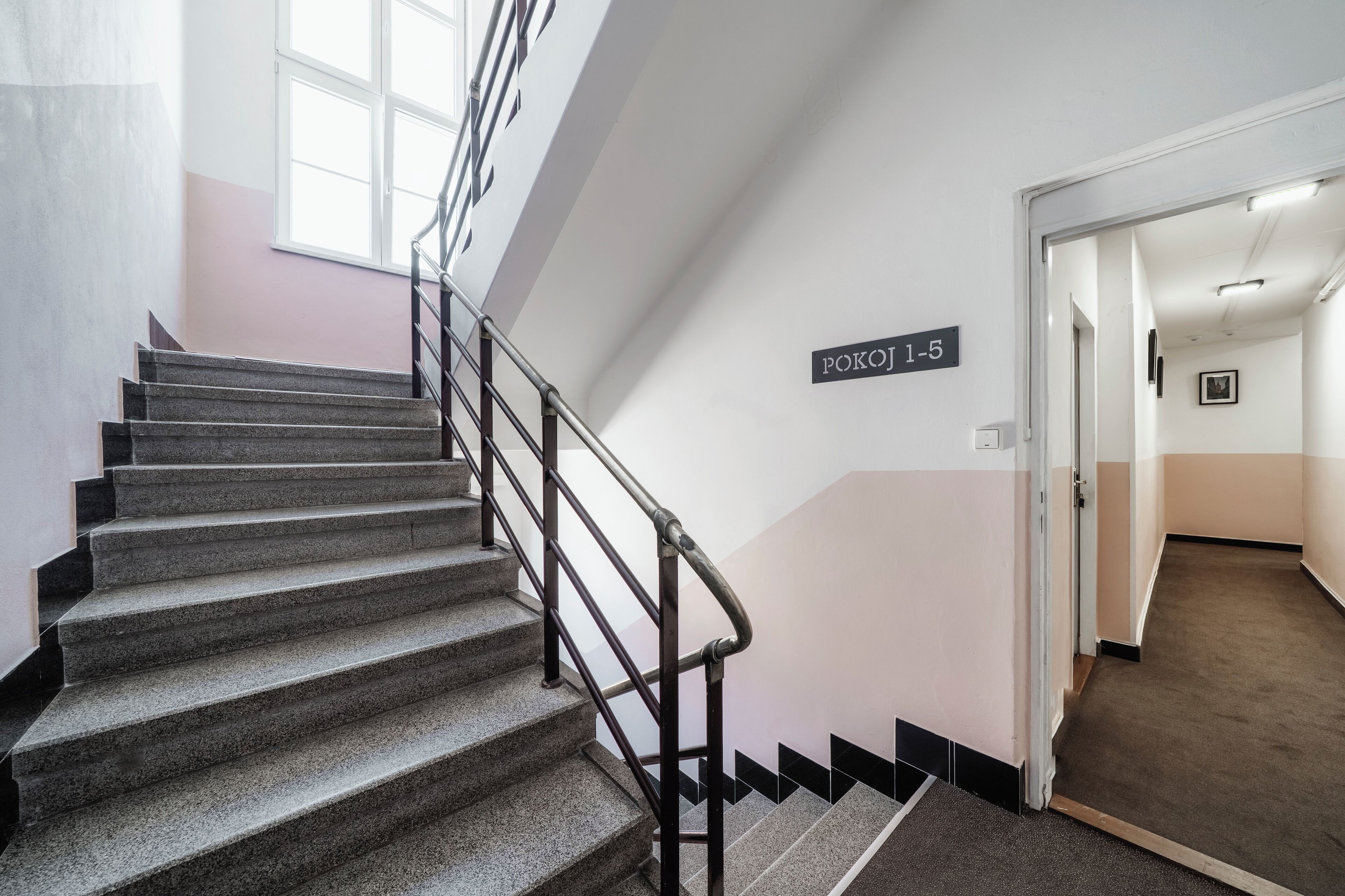 A staircase with a metal railing leading to a landing. A sign on the wall reads "POKOJI 1-6".