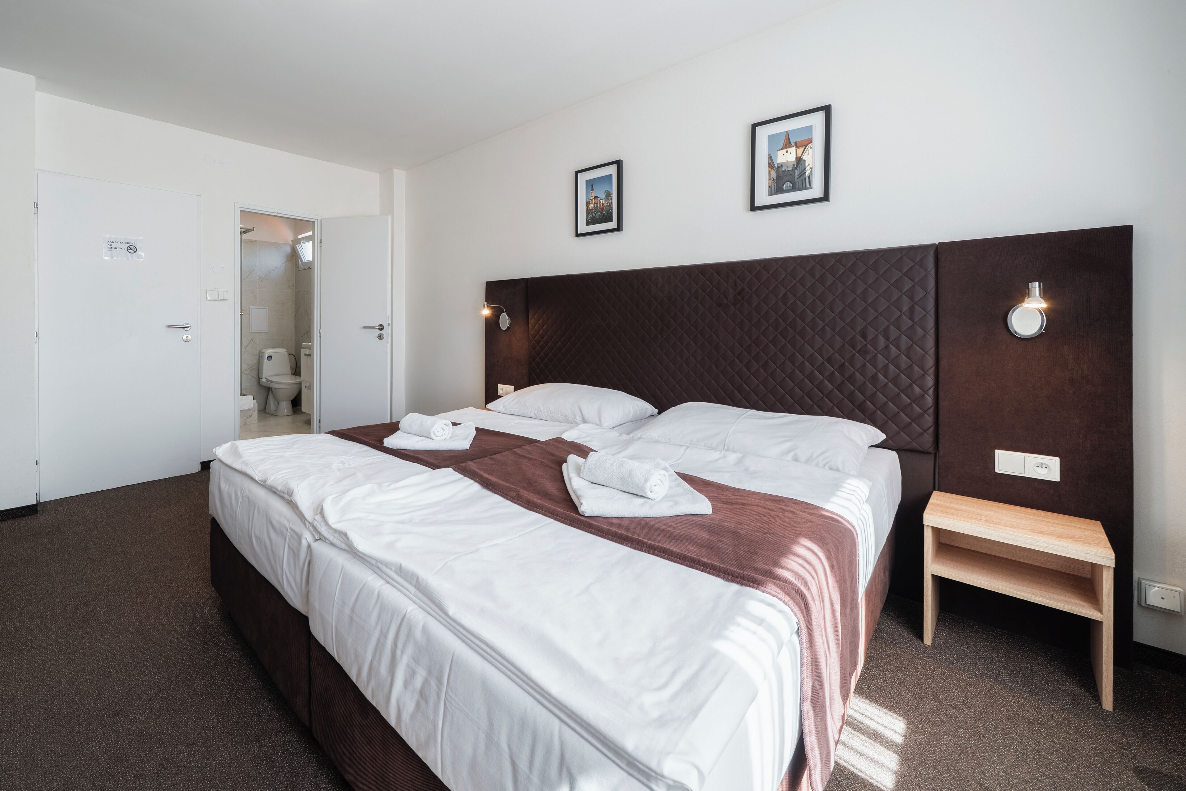 Una habitación de hotel con una cama grande, una mesita de noche de madera y dos cuadros en la pared.