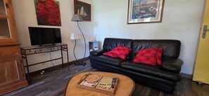 Un salon avec un canapé en cuir noir, des coussins rouges, une table basse en bois, un téléviseur posé sur un meuble et des œuvres d’art accrochées aux murs.