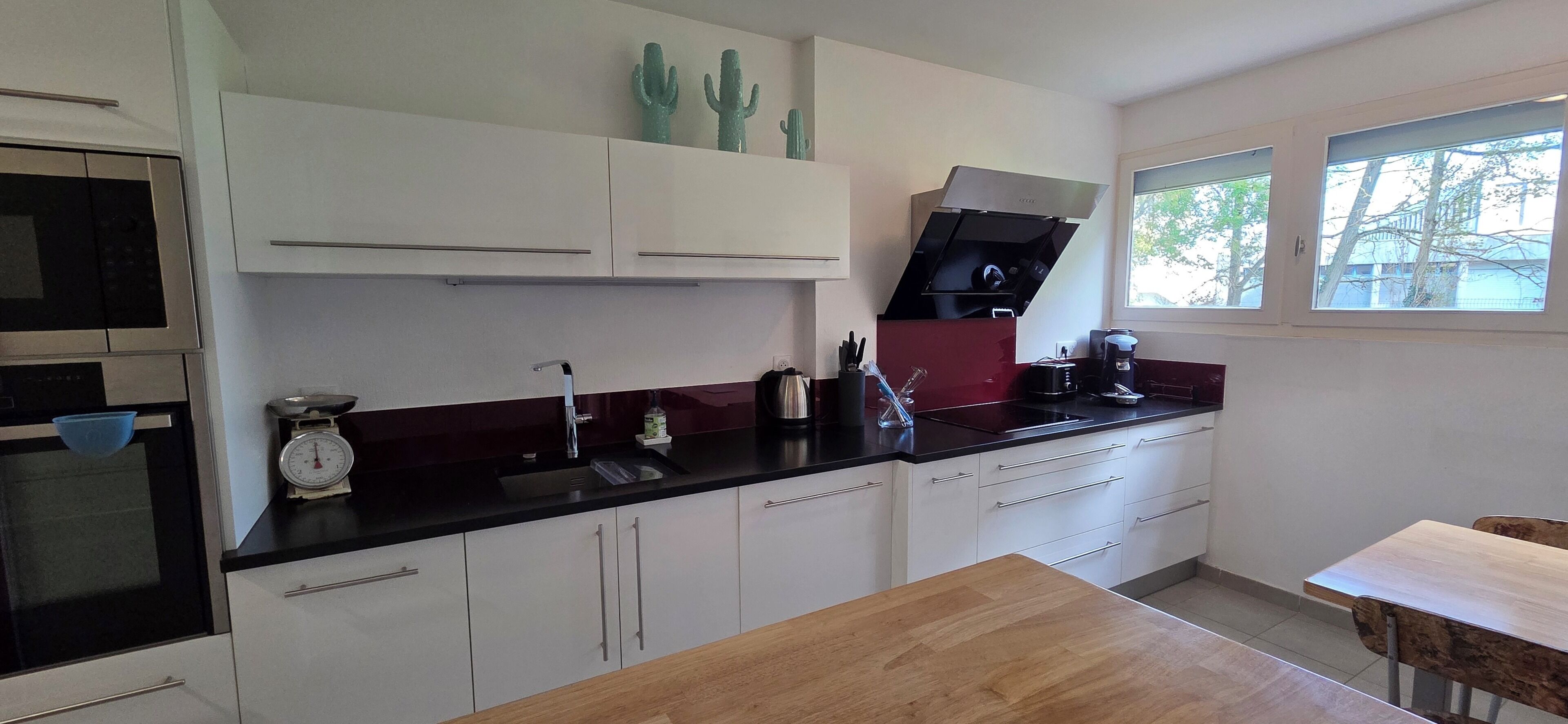 Une cuisine moderne avec des meubles blancs, un plan de travail noir, une table en bois et une fenêtre donnant sur des arbres.