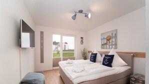 Un dormitorio con cama, una banca, televisor y ventana con vista a edificios.