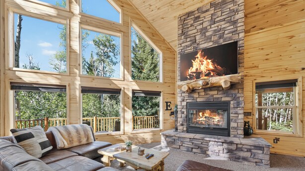 L’intérieur d’une cabane en rondins, avec une cheminée en pierre, de grandes fenêtres, un canapé en cuir et une terrasse en bois.