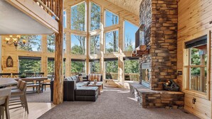 Un intérieur spacieux comprenant un grand salon, une cheminée en pierre et de nombreuses fenêtres offrant une vue sur la forêt environnante.