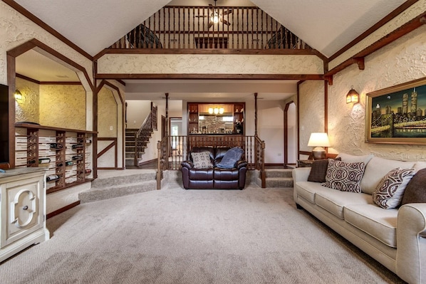 A spacious living room with a high ceiling, a staircase, a fireplace, and a wine rack.