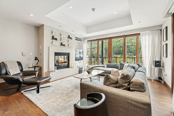 A modern living room with a fireplace, leather furniture, and large windows offering a view of the outdoors. - Private Ritz-Carlton by InvitedHome | Patio, Hike, Pool, Hot Tub, Golf Access (Vail)