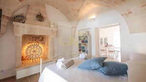 A bedroom with a stone fireplace, a bed with blue pillows, and a view of a dining area. - TD Villa Ricchiari  | Design Villa with Heated Pool (Fasano)