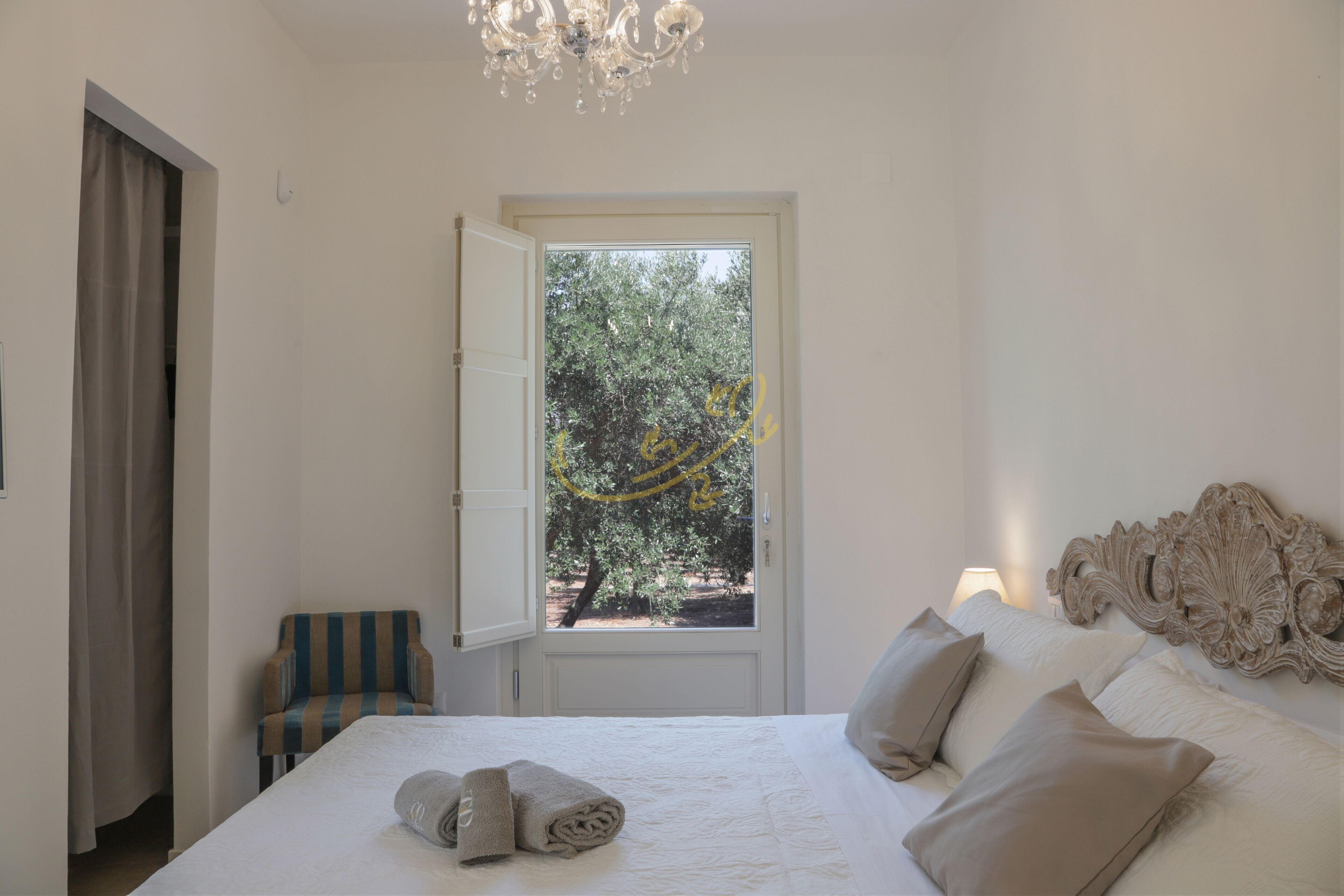 Ein Schlafzimmer mit einem großen Bett, einem Sessel, einer Kandelaberleuchte und einer Tür zu einem Außenbereich mit Bäumen.