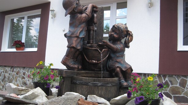 Una estatua de bronce muestra a un hombre y un niño sacando agua de un pozo, rodeados de flores y rocas.