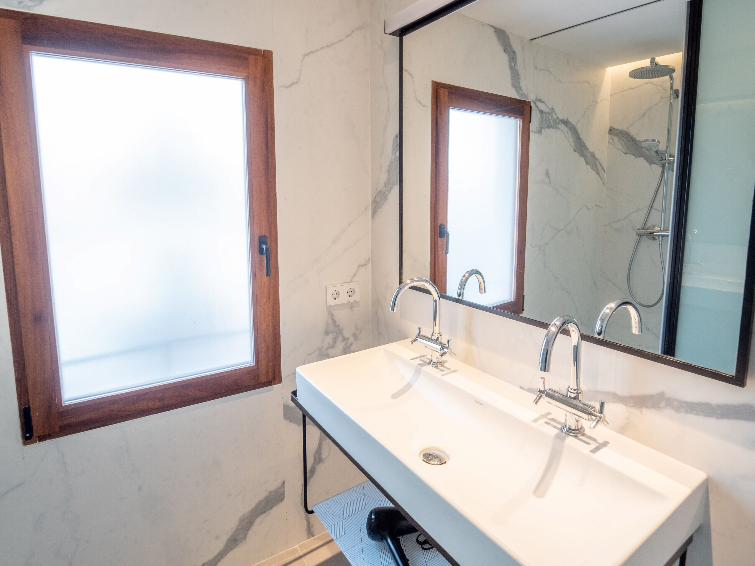 Un baño con lavamanos blanco, dos grifos, un espejo y una ducha.