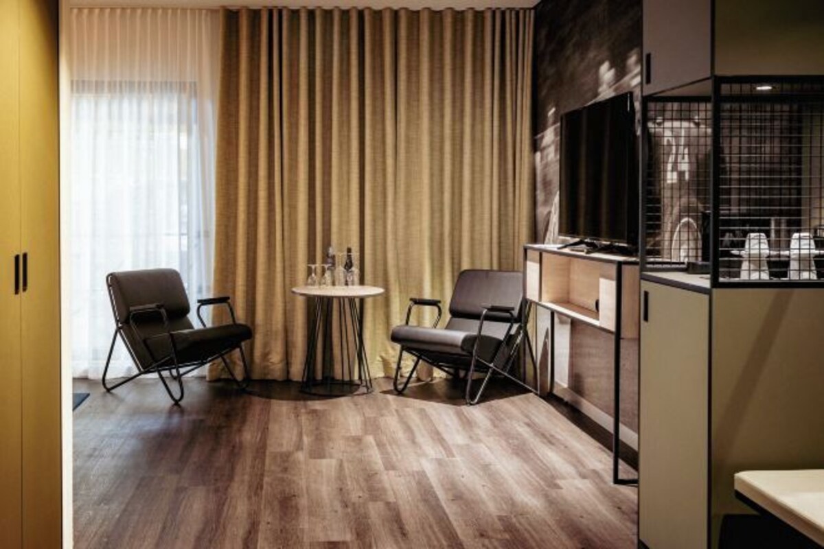 A modern hotel room with a wooden floor, two black chairs, a small round table, a large window with beige curtains, and a TV on a stand.