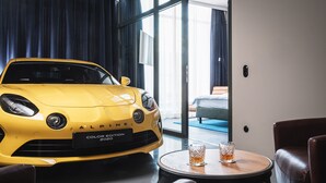 Un auto amarillo estacionado dentro de una habitación de hotel moderna, con un ventanal grande, una mesa redonda con dos vasos y muebles de cuero.