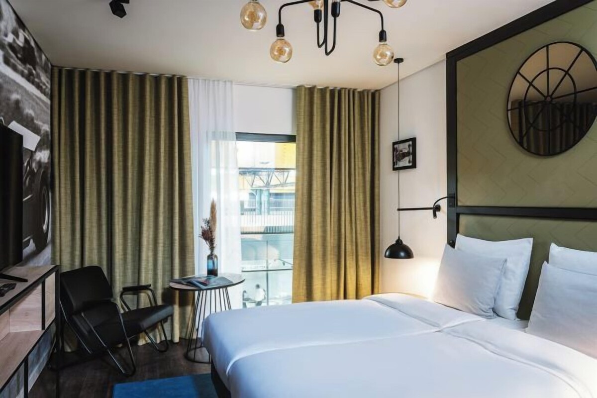 A hotel room with a bed, a desk, a chair, a lamp, and a view of the outside.