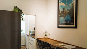 Een compacte keuken met een magnetron, een kleine koelkast en een potplant. Er staat een houten tafel met stoelen en een lijst met een afbeelding aan de muur.