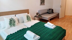 Een keurig ingerichte hotelkamer met een bed, een groene sprei, witte kussens, een tafeltje met een plant, een grijze bank en een deur.