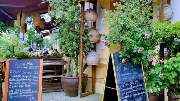 Een terras met een houten bord waarop 'gate 157' staat. Er staan potten met planten en bloemen rondom, en een krijtbordmenu is zichtbaar.