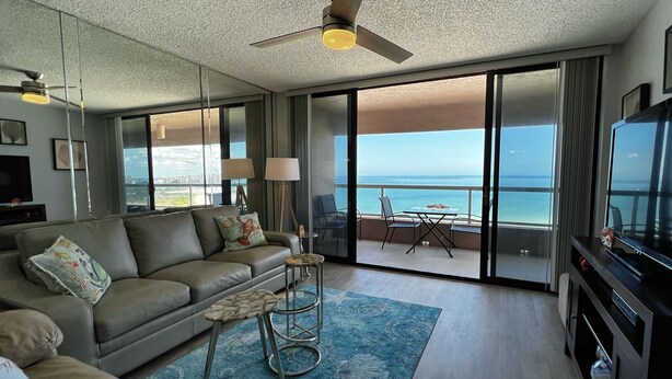 A modern living room with a sofa, a coffee table, and a TV. There is a balcony with a view of the ocean. - Beachfront Sky High Views-14th Floor- Sand Key! Resort Level Amenities! (Clearwater)