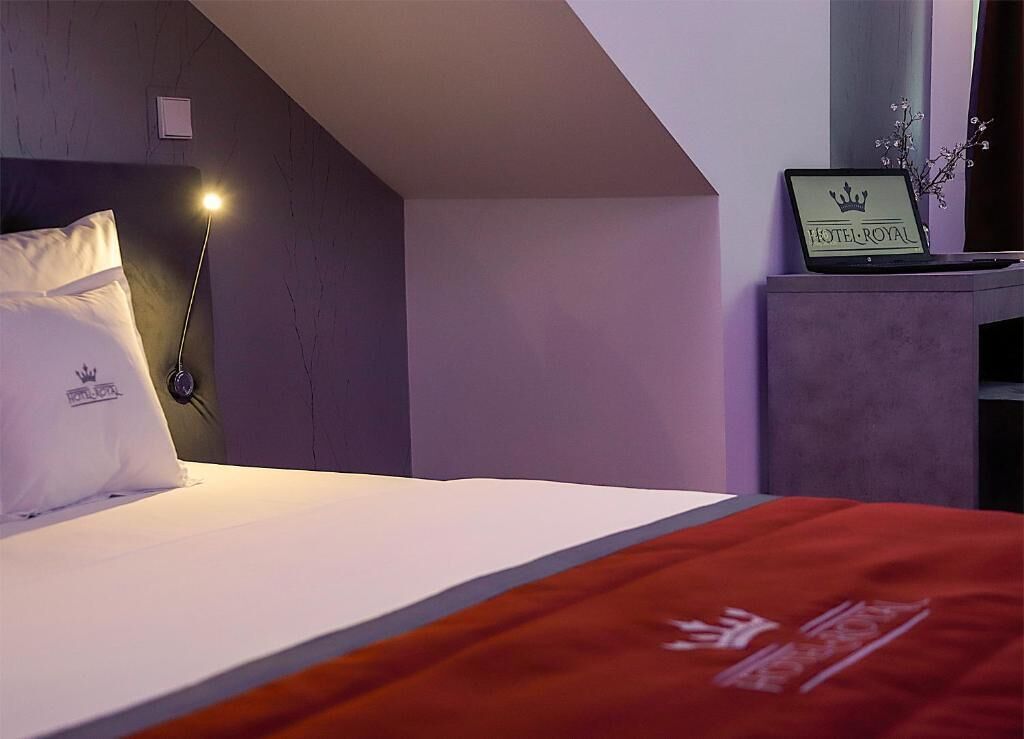 A hotel room with a bed, a nightstand, a lamp, a sign with 'HOTEL ROYAL' on it, and a window.