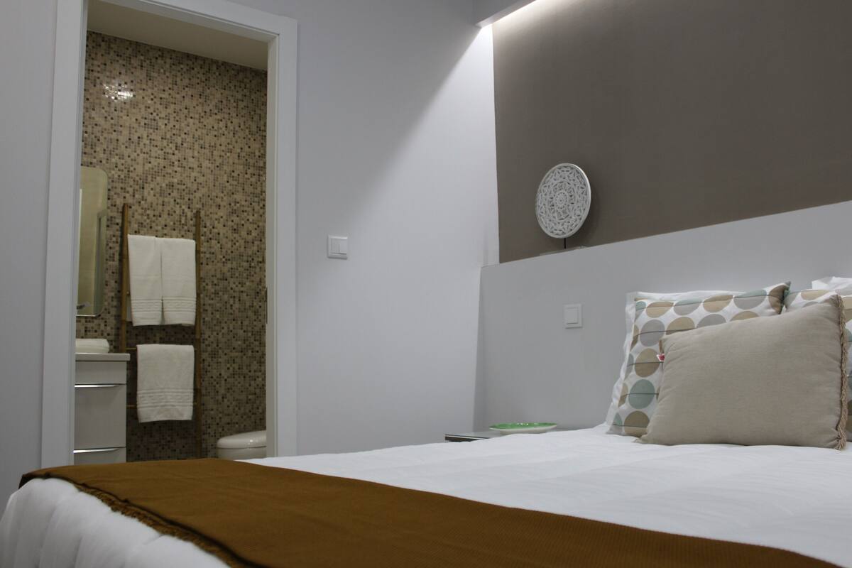 A bedroom with a bed, a bedside table, a wall-mounted decorative plate, and a bathroom visible through an open door.