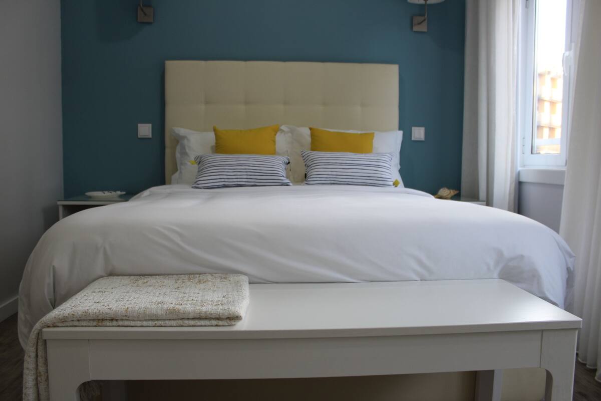 A neatly made bed with white bedding and two yellow pillows, a bedside table with a folded towel, and a nightstand with a lamp.