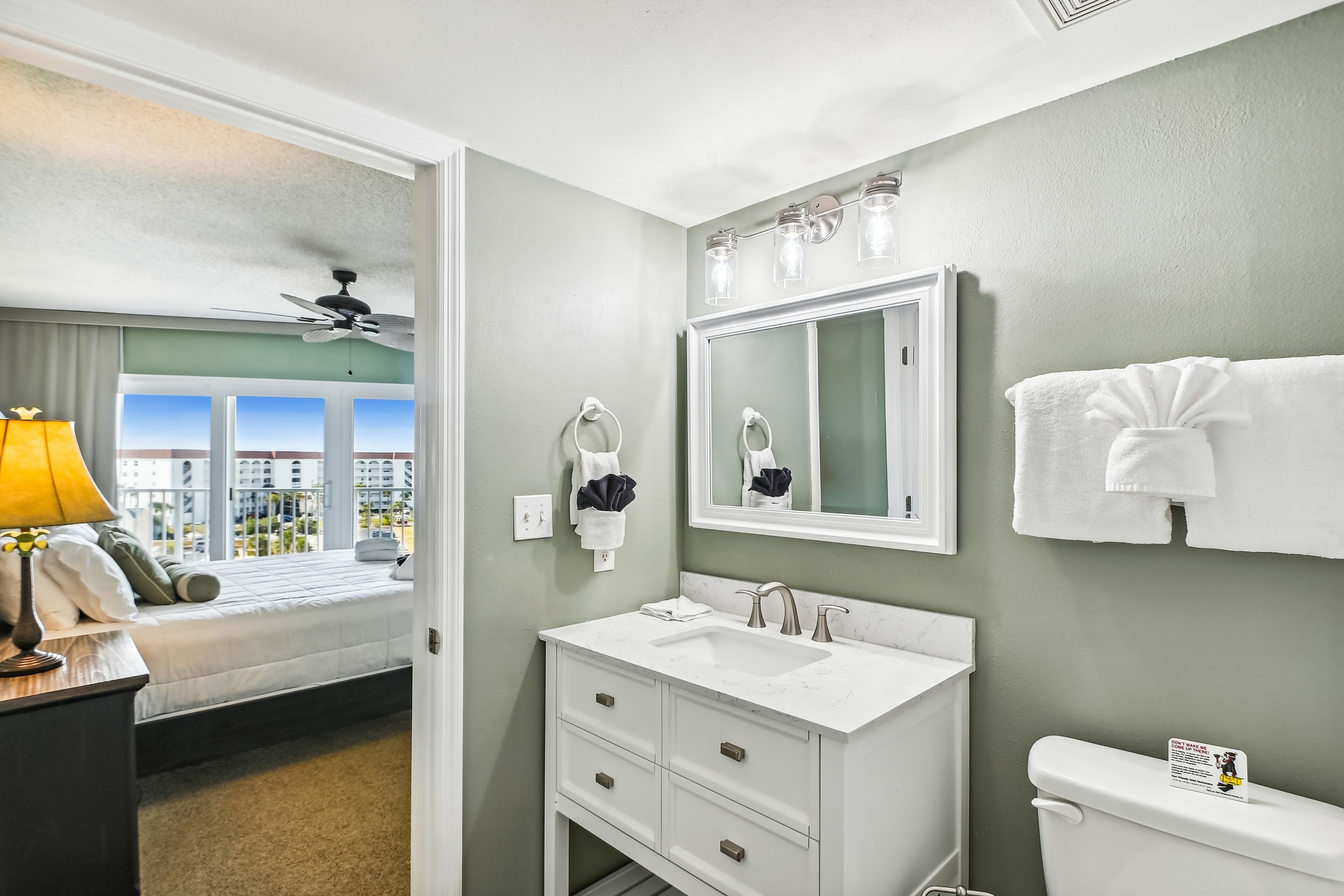 Une salle de bain d’hôtel avec une grande fenêtre, un ventilateur de plafond, un lavabo blanc et des toilettes.