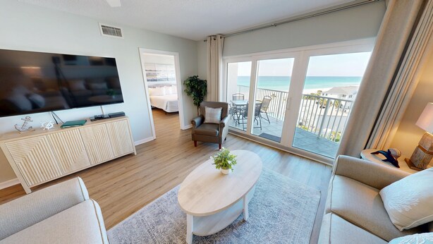 A modern living room with a flat-screen TV, a wooden cabinet, a beige sofa, a coffee table, and a balcony with a view of the sea. - Seacrest 702 is a Gulfview 2 BR on Okaloosa Island by RedAwning (Fort Walton Beach)