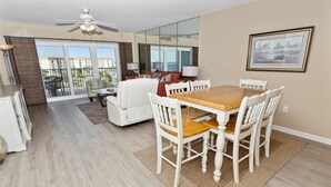 A dining area with a wooden table and white chairs, a white sofa, and a glass door leading to a balcony. - Seacrest 604 is a 2 BR Gulf Front on Okaloosa Island by RedAwning (Fort Walton Beach)