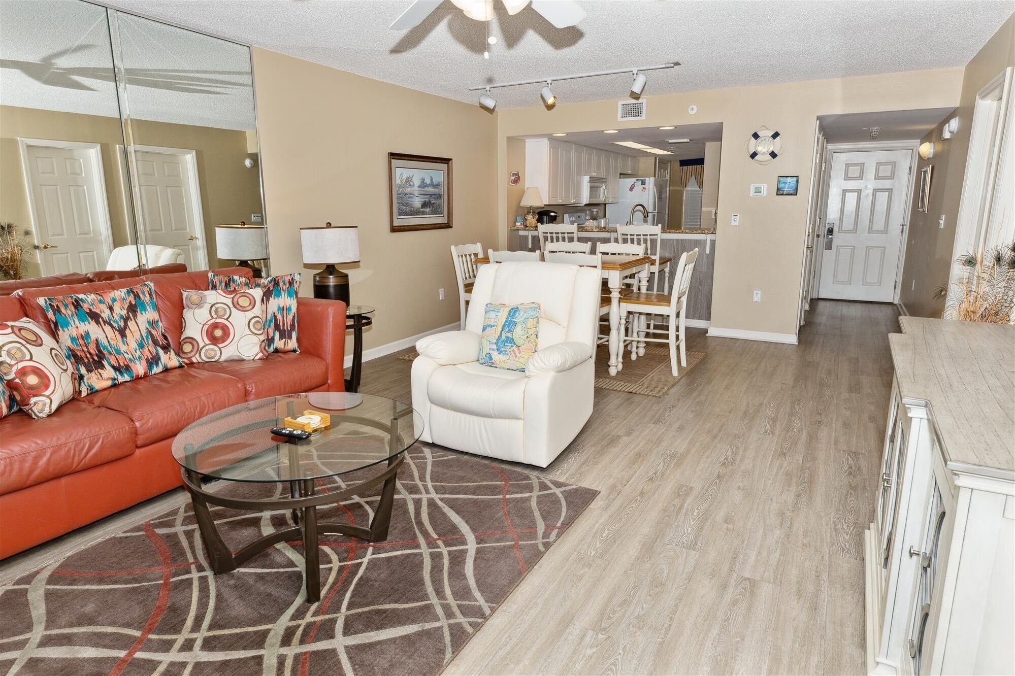 A living room with a red sofa, white armchair, and glass coffee table. There is a kitchen in the background with white cabinets and a dining area.