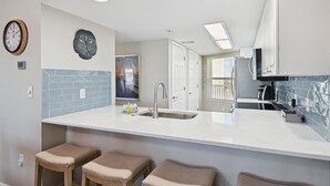 Une cuisine moderne avec un plan de travail blanc, deux tabourets de bar, un évier et une horloge au mur.