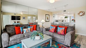 A living room with two grey sofas, colorful cushions, a white coffee table, and a large mirror. - Seacrest 511 is a Gulf side 2 BR on Okaloosa Island by RedAwning (Fort Walton Beach)