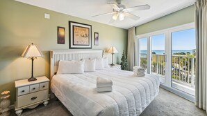 Une chambre à coucher comprenant un lit, deux tables de chevet, un ventilateur de plafond et une grande fenêtre donnant sur l’océan.