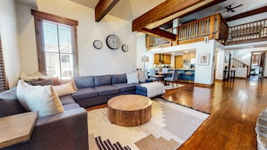 Un salón espacioso con un sofá modular gris, una mesa de centro de madera y una alfombra grande. Se ve una cocina al fondo, con armarios de madera y una zona de comedor.