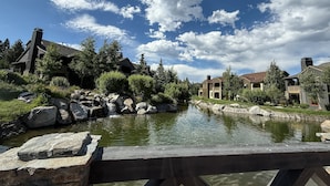 Vista a la orilla del río con un puente de madera, rocas de piedra y casas de estilo arquitectónico rústico.