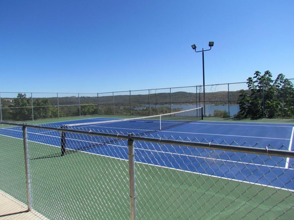 Ein Tennisplatz mit einem Drahtzaun, einer Flutlichtanlage und Hügeln sowie Wasser im Hintergrund.