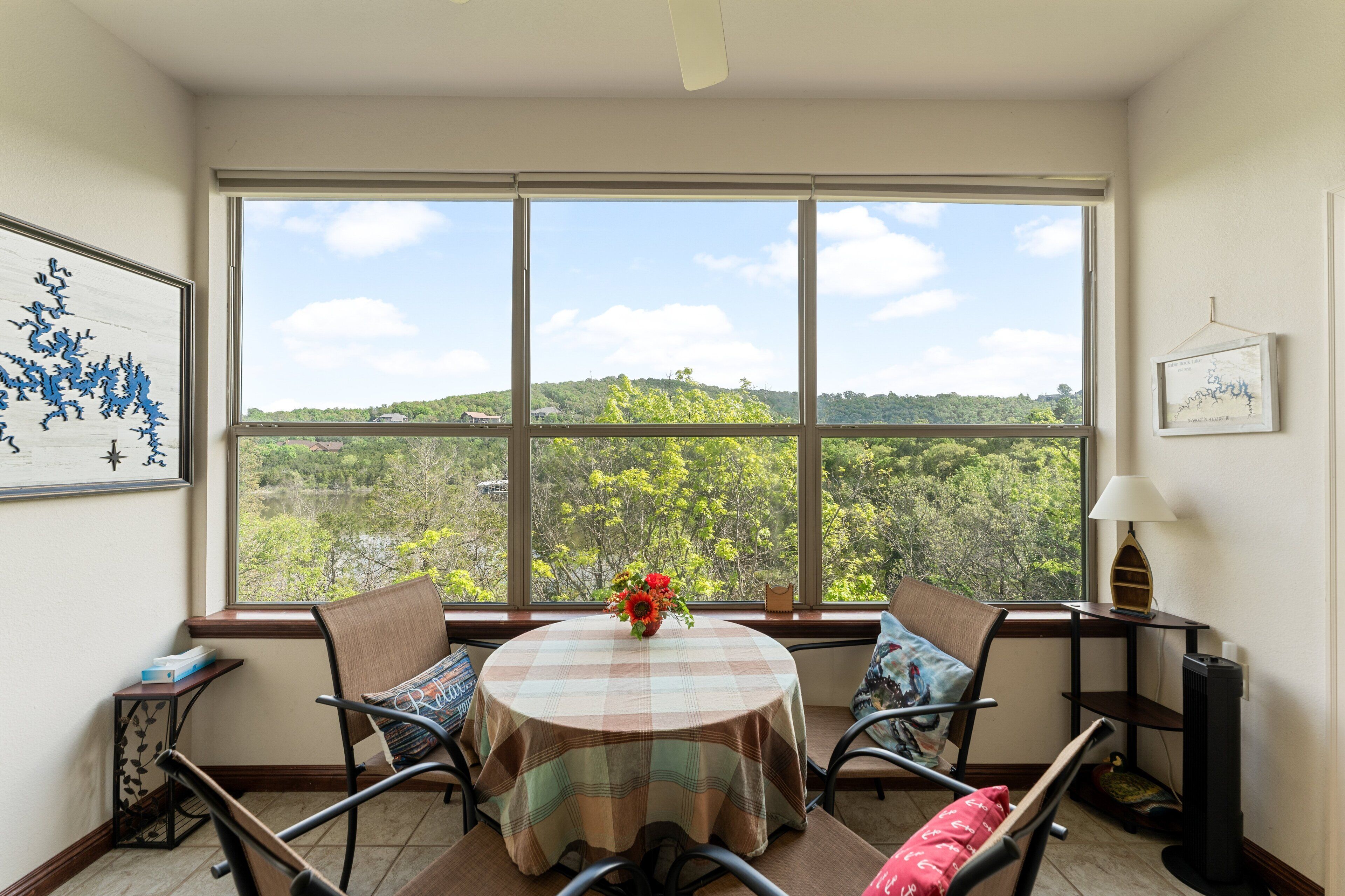A dining area with a table set for four, a window with a view of greenery, and a framed picture on the wall.