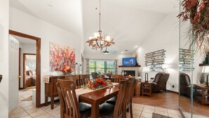 A dining area with a wooden table, chairs, and a chandelier. There is a fireplace and a large window in the background.