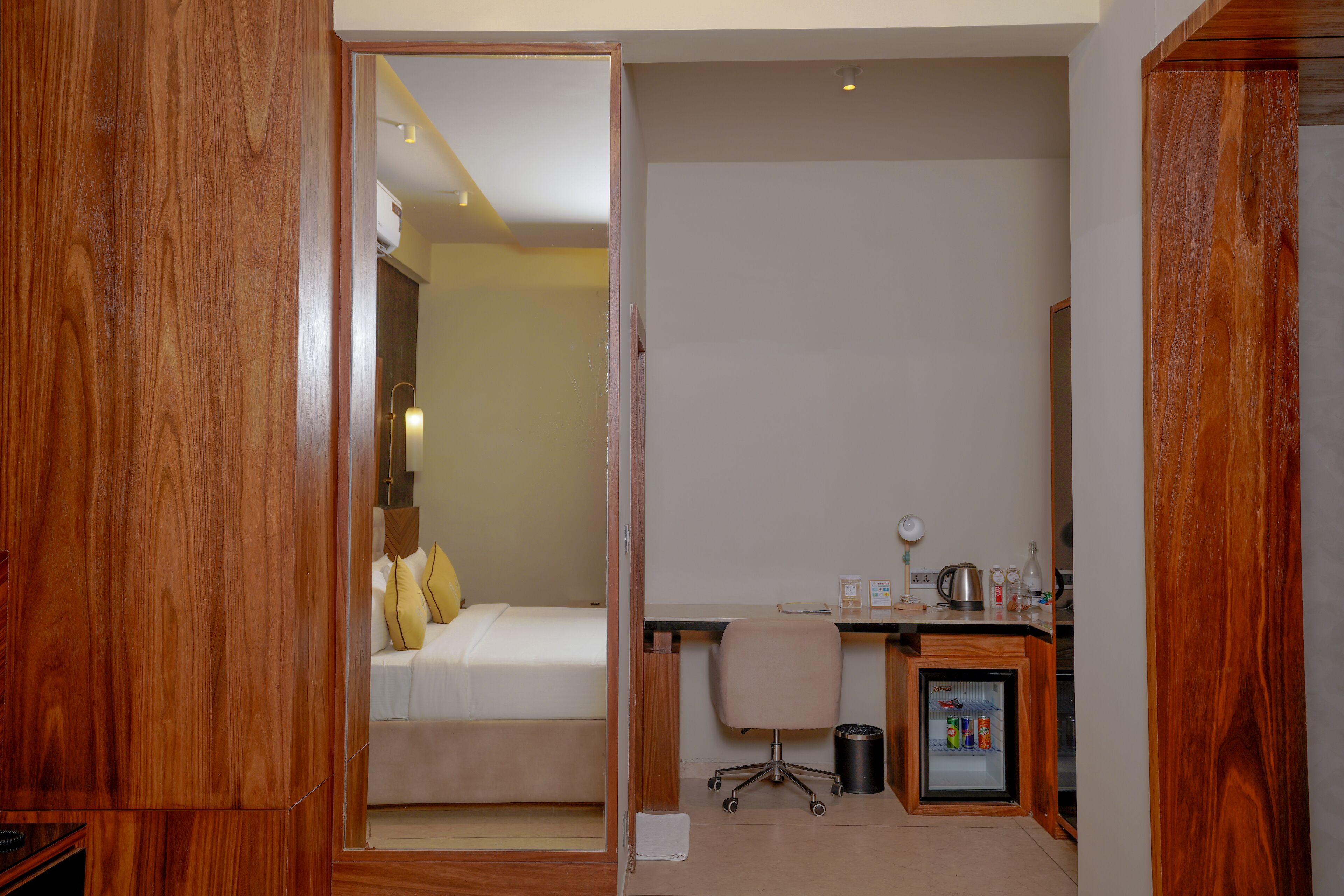 Habitación de hotel con un armario de madera, una cama con almohadas amarillas, un escritorio con una silla, un refrigerador pequeño y un cesto de basura.