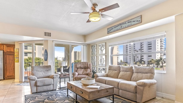 Ein großzügiges Wohnzimmer mit einem beigefarbenen Sofa, einem Relaxsessel und einem Couchtisch. Es gibt einen Deckenventilator, ein großes Fenster mit Blick auf Gebäude und ein Schild mit der Aufschrift 'New Smyrna Beach.'