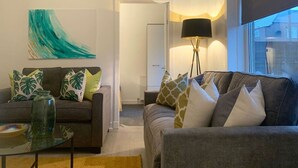 A living room with two sofas, a coffee table, and a floor lamp. There is a painting on the wall and a window with a view of buildings. - Hillington Central - stylish 3 bedroom apartment (Glasgow)