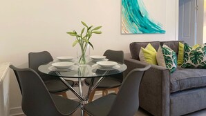 A dining area with a glass table, black chairs, and a vase with green leaves. There is a grey sofa with patterned cushions in the background. - Hillington Central - stylish 3 bedroom apartment (Glasgow)