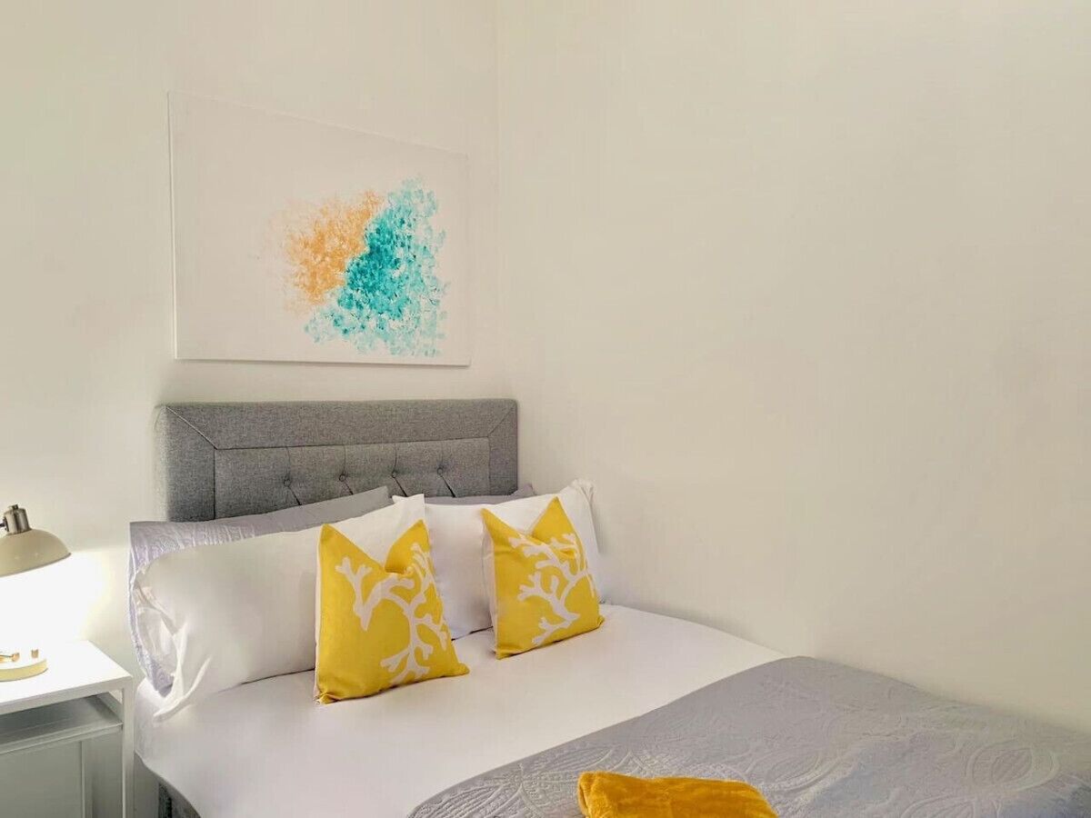 Een slaapkamer met een grijs hoofdeinde, witte beddengoed en twee gele kussens met een boommotief.