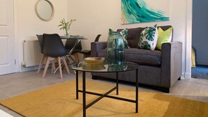 A modern living room with a glass coffee table, a grey sofa with green and yellow cushions, a round mirror on the wall, and a large abstract painting. - Hillington Central - stylish 3 bedroom apartment (Glasgow)