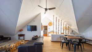 Un moderno loft con soffitto alto, ventilatore a soffitto, zona pranzo con tavolo e sedie in legno, divano in pelle nera e televisore a parete.