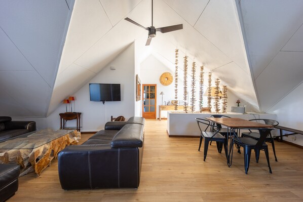 Un loft moderno con techos altos, ventilador de techo, comedor con mesa y sillas de madera, sofá de cuero negro y televisor montado en la pared.