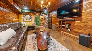 A cozy log cabin interior with a leather sofa, a rustic coffee table, and a flat-screen TV mounted on the wall. - The Hobbit House, Fantasy comes Home! (Russellville)