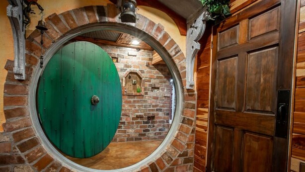 A circular door with a green surface, a brick wall with a small window, and a wooden door to the right. - The Hobbit House, Fantasy comes Home! (Russellville)
