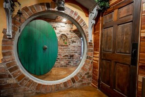 Une porte circulaire à la surface verte, un mur de briques avec une petite fenêtre et une porte en bois sur la droite.