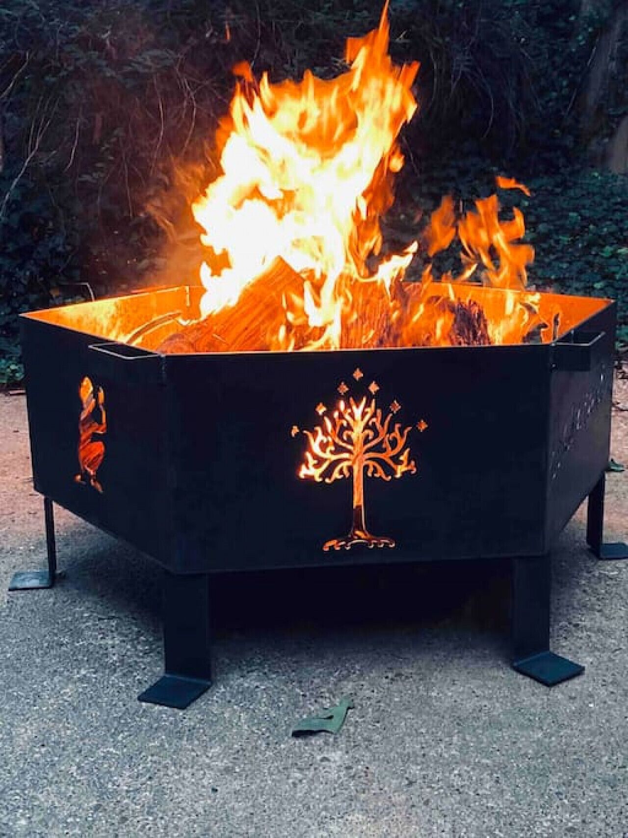 A black outdoor fire pit with a tree design, surrounded by gravel.