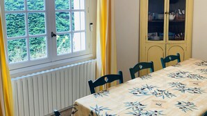 Ruokailuhuone, jossa on neljälle katettu ruokapöytä, ikkuna verhoilla, patteri ja lasiovilla varustettu kaappi.