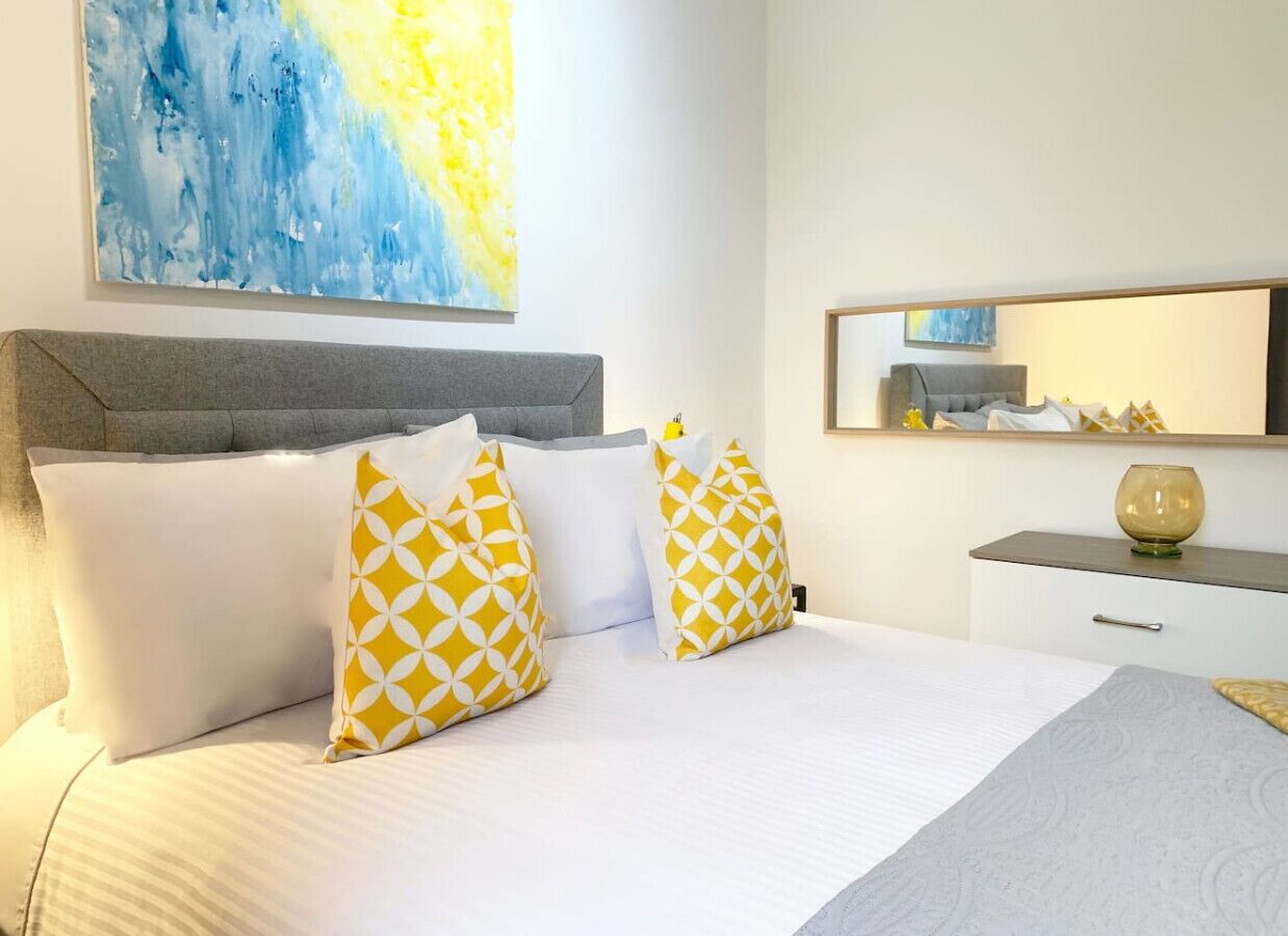 A hotel room with a bed, two pillows, a bedside table, a lamp, a mirror, and a painting on the wall.