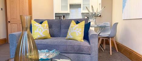 Ein Wohnzimmer mit grauem Sofa, gelb-blauen Kissen mit Muster, einem Glastisch mit einer Vase und einer dekorativen Schale, sowie einem Essbereich mit weißem Tisch und Stühlen.